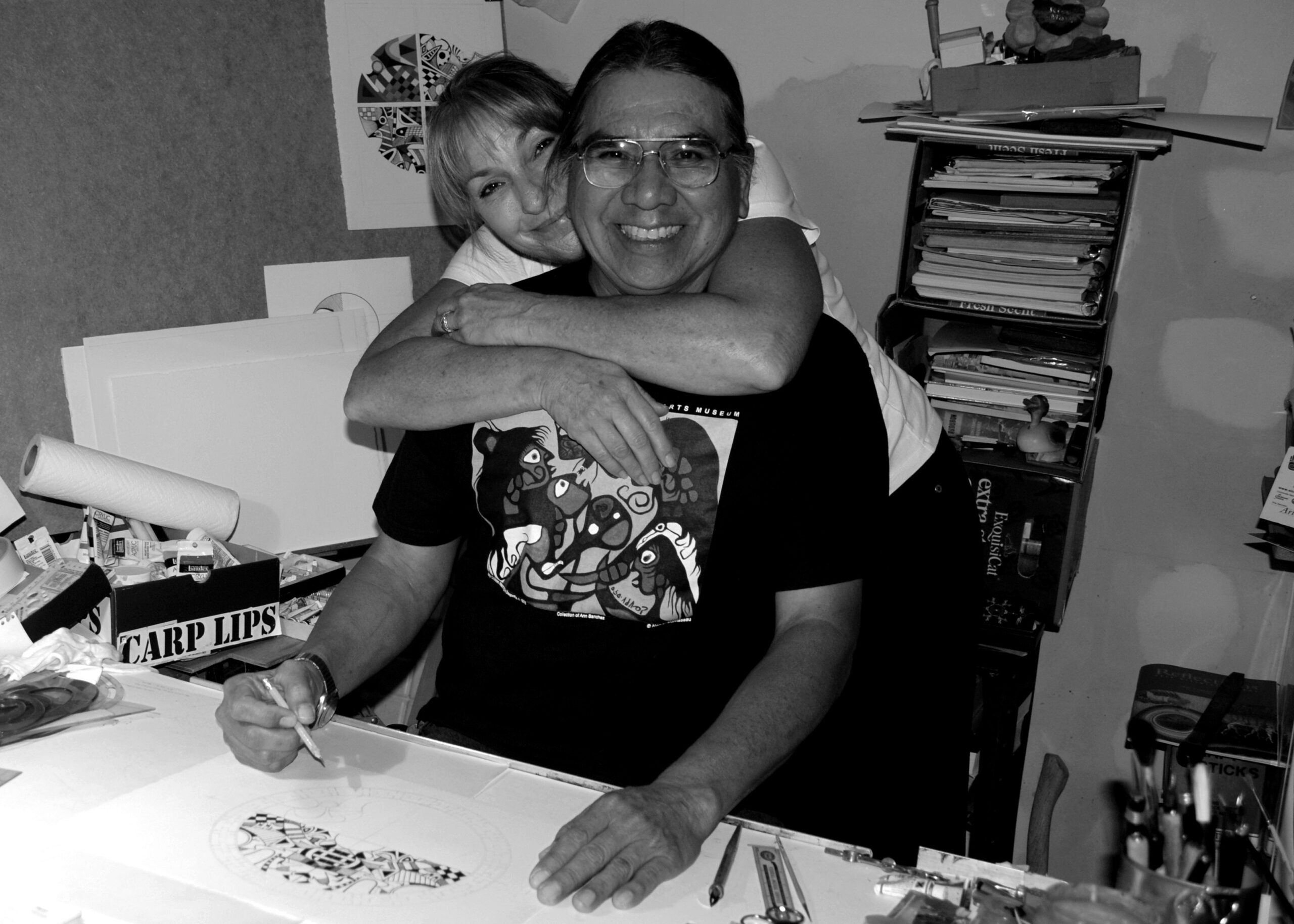 Ben seated in his studio working on a painting, smiling at the camera, with Barbara's arms wrapped around him from behind in an embrace