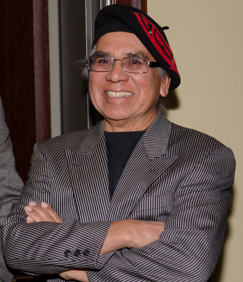 A portrait of Ben smiling wearing glasses and a black beret adorned with red and black designs, wearing a striped suit jacket, stands with his arms crossed